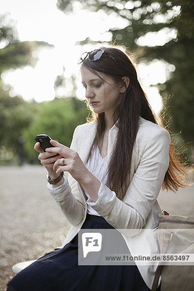 Eine Frau in einer cremefarbenen Jacke  die in einem Stadtpark sitzt und auf ihr Handy schaut.