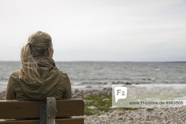 Eine Frau mit langen blonden Haaren,  die einen braunen Kapuzenmantel trägt und auf einer Bank mit Blick aufs Meer sitzt.