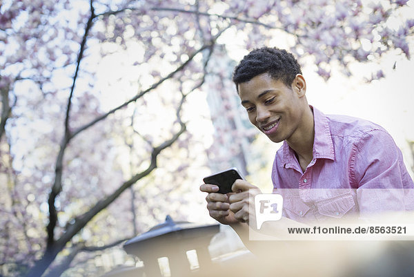 Stadtleben. Ein junger Mann im Frühling im Park  der ein Mobiltelefon benutzt.