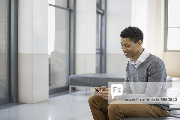 Urbaner Lebensstil. Ein junger Mann sitzt in einer Lobby  auf einer Sitzbank. Er benutzt sein Mobiltelefon.