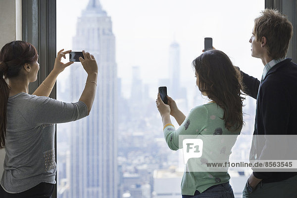 Urbaner Lebensstil. Drei Personen  die auf einer Aussichtsplattform stehen und mit ihren Telefonen Bilder von der Aussicht über die Stadt aufnehmen.