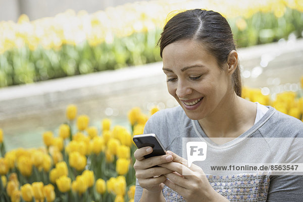 Urbaner Lebensstil. Eine Frau im Park  an einem Bett aus gelben Tulpen  die ihr Mobiltelefon benutzt.