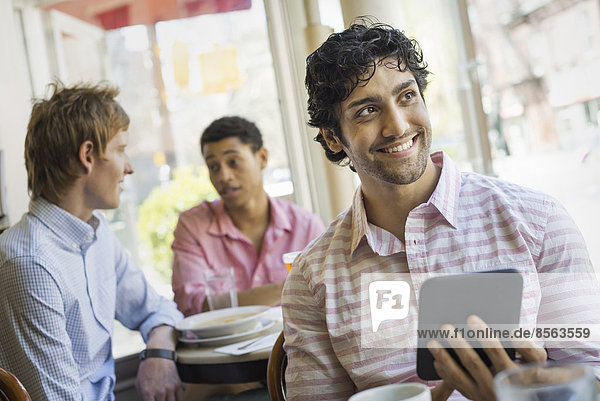 Urbaner Lebensstil. Drei junge Männer um einen Tisch in einem Cafe. Einer hält ein digitales Tablett in der Hand.