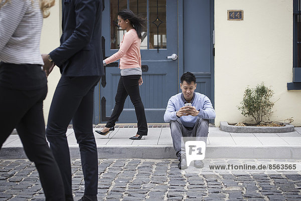Young people outdoors on the city streets in springtime. A man sitting on the ground checking his phone  and three passers-by.