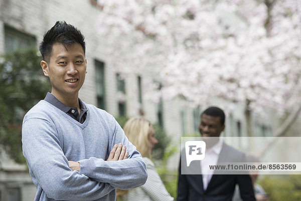 City life in spring. Young people outdoors in a city park. A man with his arms folded and two people talking in the background under the trees in blossom.
