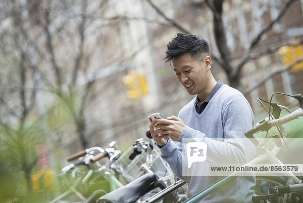 Stadtleben im Frühling. Ein Mann in einem blauen Pullover an einer Reihe geparkter Fahrräder. Seine Nachrichten auf einem Smartphone abrufen.
