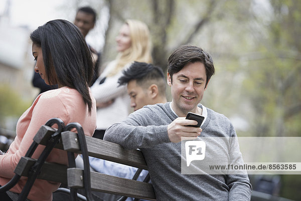 City life in spring. Young people outdoors in a city park. Sitting on a park bench. Five people  men and women  checking their phones.