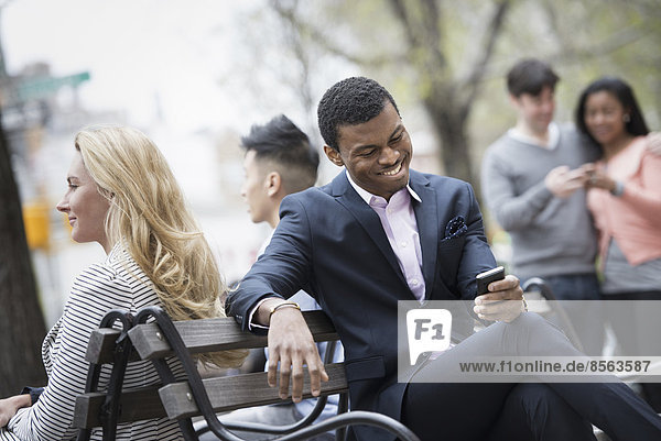 City life in spring. Young people outdoors in a city park. A group of men and women gathered around a park bench. Two checking a smart phone for messages.