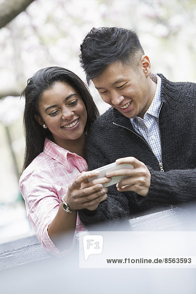 Stadtleben im Frühling. Jugendliche im Freien in einem Stadtpark. Ein Paar nebeneinander  auf ein Smartphone blickend.