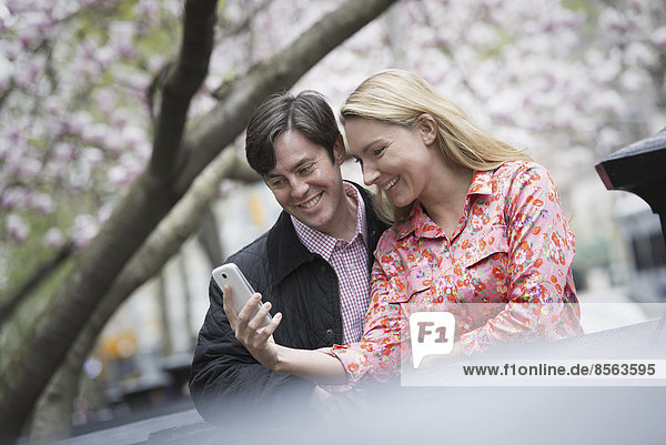 City life in spring. Young people outdoors in a city park. A young woman and man sitting side by side looking down at a smart phone.