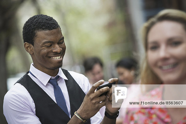 Stadtleben im Frühling. Jugendliche im Freien in einem Stadtpark. Ein Mann lächelt  als er auf sein Telefon schaut  und die Nahaufnahme einer Frau mit blonden Haaren.
