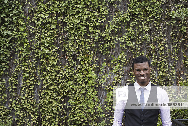 Stadtleben im Frühling. Stadtpark mit einer mit Kletterpflanzen und Efeu bewachsenen Wand. Ein junger Mann mit Weste  Hemd und Krawatte. Ein Blick in die Kamera.