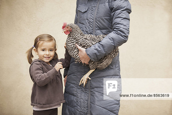Eine Frau in grauem Mantel  die ein schwarz-weißes Huhn mit rotem Hahnenkamm unter einem Arm hält. Neben ihr ein junges Mädchen  das die andere Hand hält.