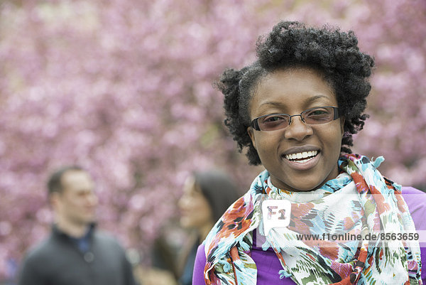 A group of people under the cherry blossom trees in the park. A young woman smiling and looking at the camera. Wearing a floral scarf.