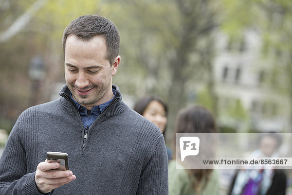 Eine Gruppe von Menschen in einem Stadtpark. Ein Mann in einem grauen Pullover  der sein Telefon überprüft.