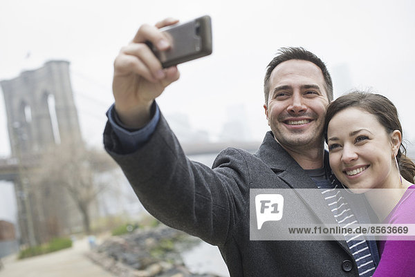 New York Stadt. Die Brooklyn Bridge über den East River. Ein Paar beim Fotografieren mit einem Telefon,  ein Selfy von sich selbst.