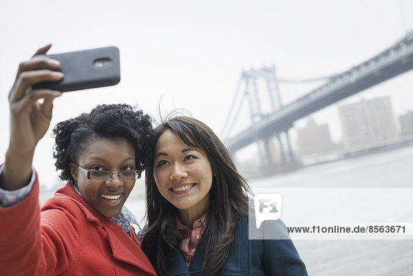 New York Stadt. Die Brooklyn Bridge über den East River. Ein Paar  zwei Frauen  die ein Foto mit einem Telefon machen  ein Selbstbild von sich selbst.