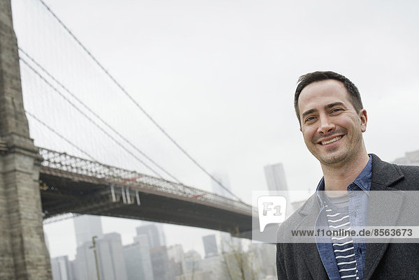 New York City  die Brooklyn Bridge  die über den East River führt. Ein Mann in einem grauen Mantel lächelt in die Kamera.