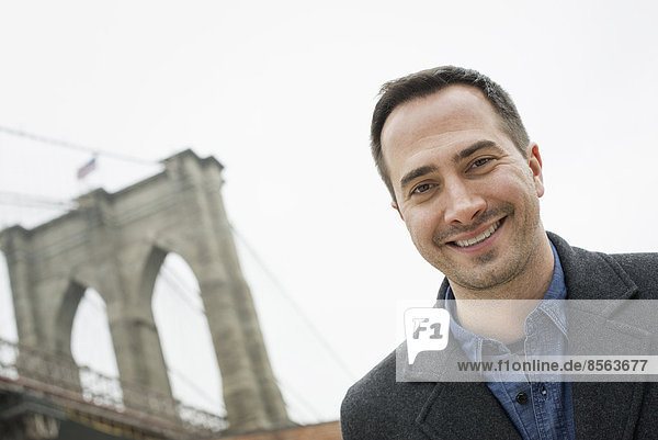 New York City  die Brooklyn Bridge  die über den East River führt. Ein Mann  der in die Kamera schaut und lächelt.