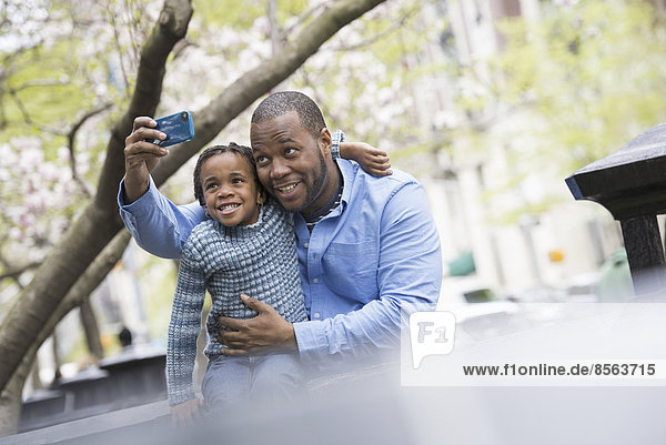 Ein New Yorker Stadtpark im Frühling. Sonnenschein und Kirschblüte. Vater und Sohn Seite an Seite. Mit einem Smartphone ein Foto machen.