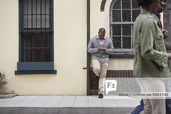 Draußen in der Stadt im Frühling. Ein Paar  das auf dem Bürgersteig geht  und ein Mann  der an einer Wand lehnt und sein Telefon überprüft.