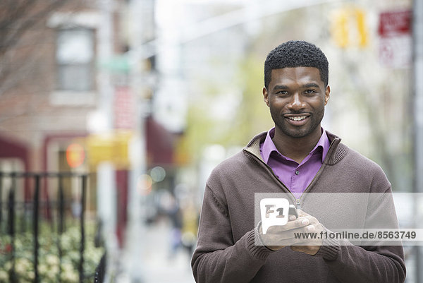 Draußen in der Stadt im Frühling. Ein urbaner Lebensstil. Ein junger Mann hält sein Telefon und lächelt in die Kamera.