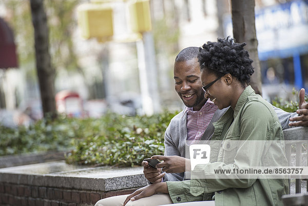 Draußen in der Stadt im Frühling. Ein urbaner Lebensstil. Ein Paar  das nebeneinander sitzt und auf einen Telefonbildschirm schaut.