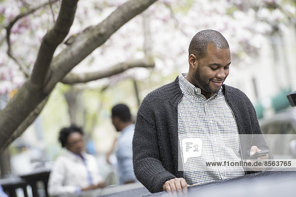 Draußen in der Stadt im Frühling. Ein urbaner Lebensstil. Ein junger Mann  der sein Telefon überprüft und SMS schreibt. Ein Paar im Hintergrund.