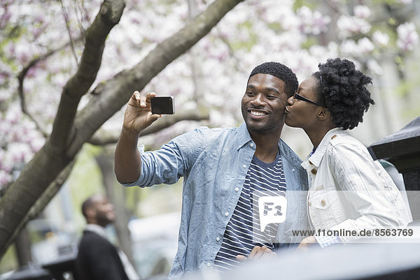 Draußen in der Stadt im Frühling. Ein urbaner Lebensstil. Eine Frau  die einen Mann küsst und mit einem tragbaren Mobiltelefon fotografiert.
