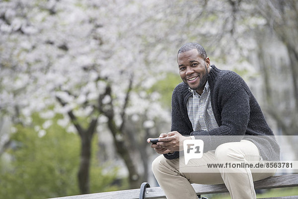 Draußen in der Stadt im Frühling. Ein urbaner Lebensstil. Ein Mann  der im Park auf einer Bank sitzt und ein Telefon hält.