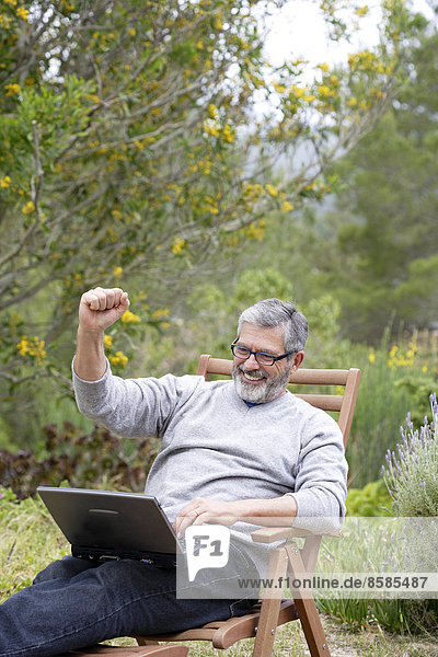 Man sitting in his garden with laptop  smiling  wearing glasses