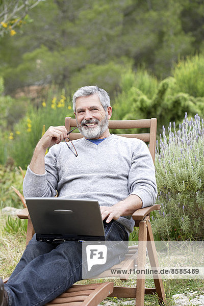 Mann sitzt in seinem Garten mit Laptop  lächelnd