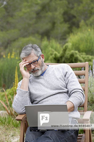 Man sitting in his garden  reading emails on his laptop
