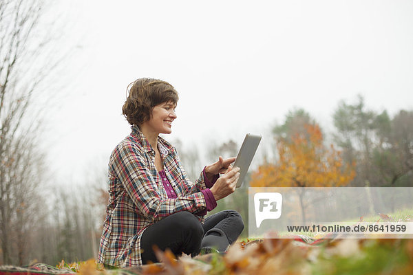 A woman using a computer tablet sitting on the ground. Autumn leaves and woodland.