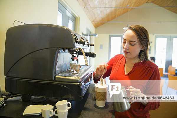 A young woman making coffee using a large coffee machine. Pouring frothed milk onto a cup.
