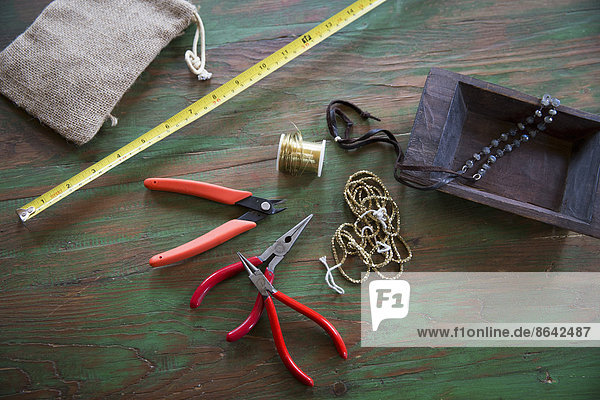 A tabletop with jewellery making equipment. Pliers  thread and a wooden tray  with measuring tape.