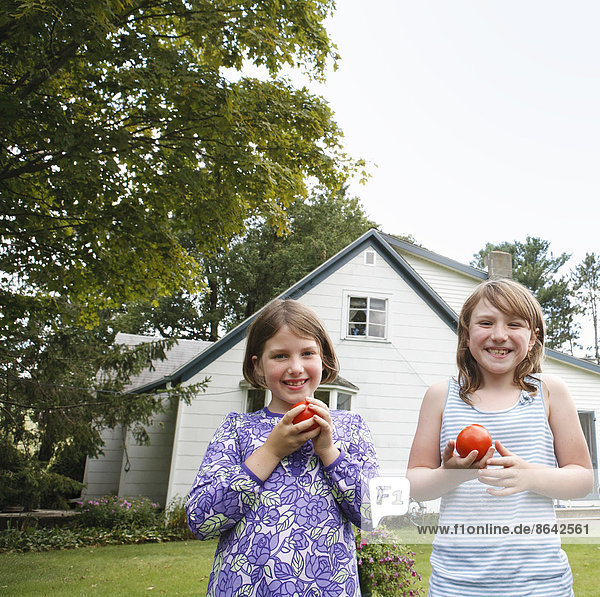 Zwei Kinder  Mädchen  die in einem Garten stehen und frisch gepflückte Tomaten halten und essen.