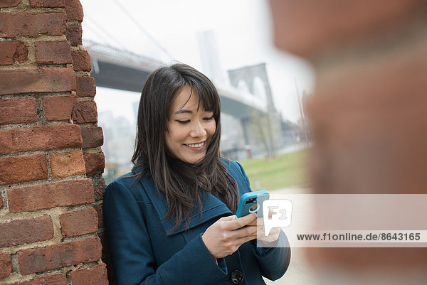 New York Stadt. Die Brooklyn Bridge über den East River. Eine Frau lehnt an einer Ziegelmauer und überprüft ihr Telefon.