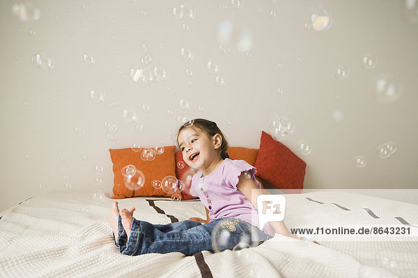 Ein junges Mädchen mit braunen Augen und dunklen Haaren in Büscheln sitzt lachend auf einem Bett. Luftblasen schweben in der Luft.