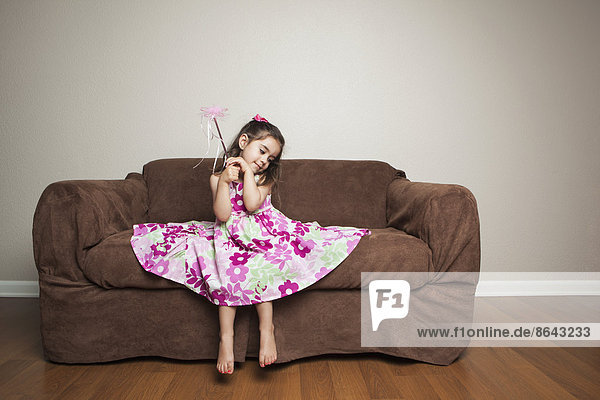 A 3 year old girl with long brown hair in a pink flowered cotton dress with the skirt spread out  waving a wand.