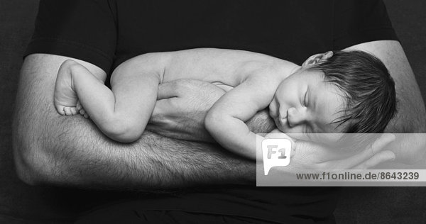 A naked newborn baby sleeping in his father's arms.