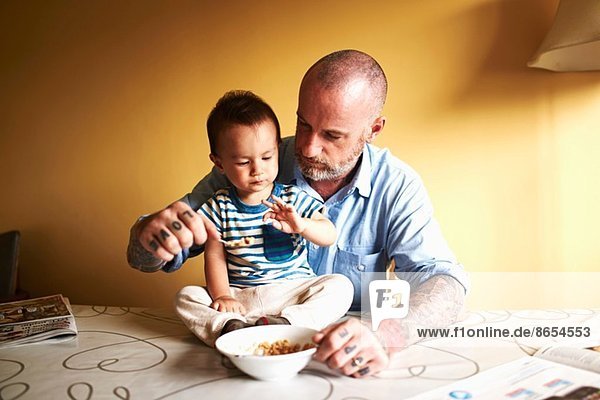 Kleiner Junge sitzt auf dem Tisch und trinkt Müsli mit Vater.