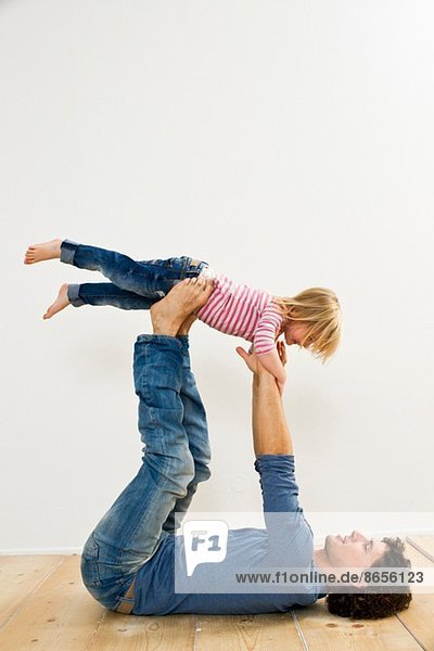 Studioaufnahme eines Vaters  der seine Tochter auf dem Rücken liegend hochhebt.