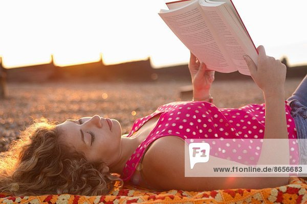 Young woman lying on beach reading book