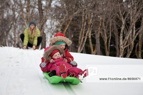 Mutter beobachtet zwei junge Töchter beim Schlittenfahren im Schnee