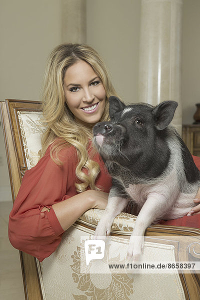 A miniature pot bellied pig sitting on his haunches on the lap of a blonde haired woman  sharing an antique armchair with thick cushions  in a large elegantly furnished mansion in Texas.