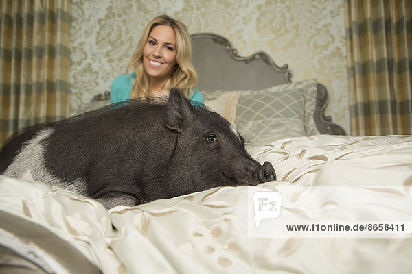 Ein Hängebauchschwein,  das auf einem großen Bett mit geschnitztem Kopfteil und Kissen liegt,  in einem großen Herrenhaus,  neben dem Besitzer,  einer Frau mit blonden Haaren.