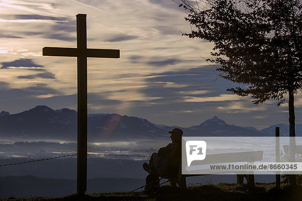 Wanderer auf Bank neben Kreuz  mit Alpenpanorama  Auerberg  Ostallgäu  Bayern  Deutschland