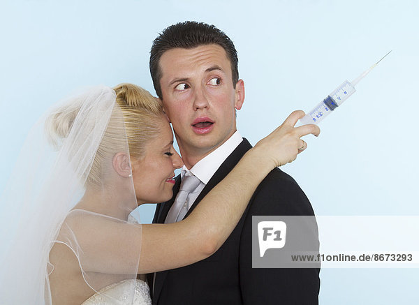 Bräutigam schaut verwundert über seine Schulter auf eine Spritze in der Hand seiner lächelnden Braut  Deutschland