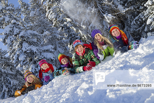 Jugendliche in bunter Winterkleidung liegen fröhlich nebeneinander im Schnee  hinten ein verschneiter Nadelwald  Tirol  Österreich